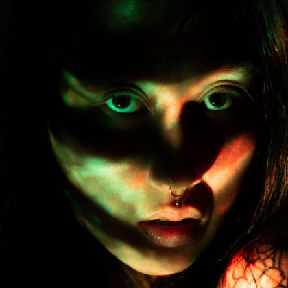 A self portrait photographs of Elaine. Her expression is neutral. Her face is slightly lit up with green and red lighting, however, there are some mysterious shadows casted over her face. The shadows look a bit like a mask over the eyes. She’s looking directly into the lens and both eyes are visible.