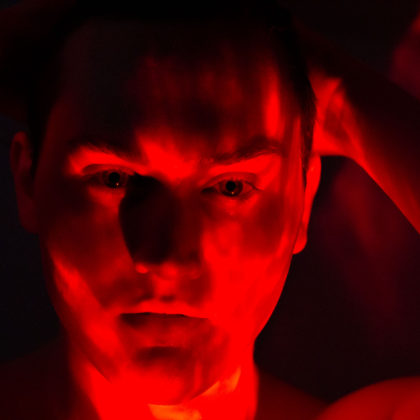 A portrait photograph of Sebastian Penn. He’s in a dark room, dimly lit with red lighting from below him. He’s pulling both hands through his hair and staring directly at the camera with a fairly neutral expression.