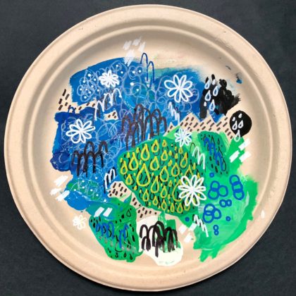 A traditional artwork of mixed media on a recycled paper plate. There are are smudges of paint – mainly in blues and greens – as well as scribbles/doodles in marker and pencil.