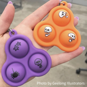 Photo of a pop-it halloween pack #1 on a hand, with an orange skelly pop-it with 3 bubbles and a purple spooky pop-it with 3 bubbles. Photo by Geelong Illustrators.