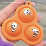 Photo of a skelly pop-it on a hand. It's orange with 3 bubbles and each bubble has a skull doodle on it. Photo by Geelong Illustrators.
