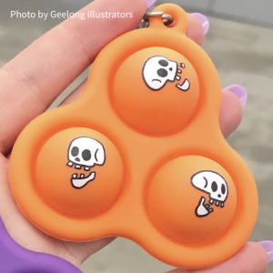Photo of a skelly pop-it on a hand. It's orange with 3 bubbles and each bubble has a skull doodle on it. Photo by Geelong Illustrators.