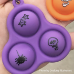 Photo of a skelly pop-it on a hand. It's purple with 3 bubbles and each bubble has a some somewhat spooky doodle illustrations on them: a skull, a spider and some stars. Photo by Geelong Illustrators.