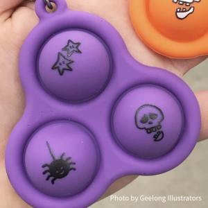 Photo of a skelly pop-it on a hand. It's purple with 3 bubbles and each bubble has a some somewhat spooky doodle illustrations on them: a skull, a spider and some stars. Photo by Geelong Illustrators.